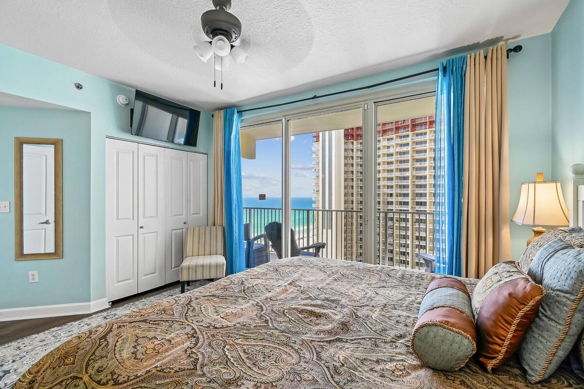 Ein Hotelzimmer mit einem großen Bett, einem Fernseher auf einer Kommode, einem Balkon mit Blick auf die Stadt und einem Sessel am Fenster.