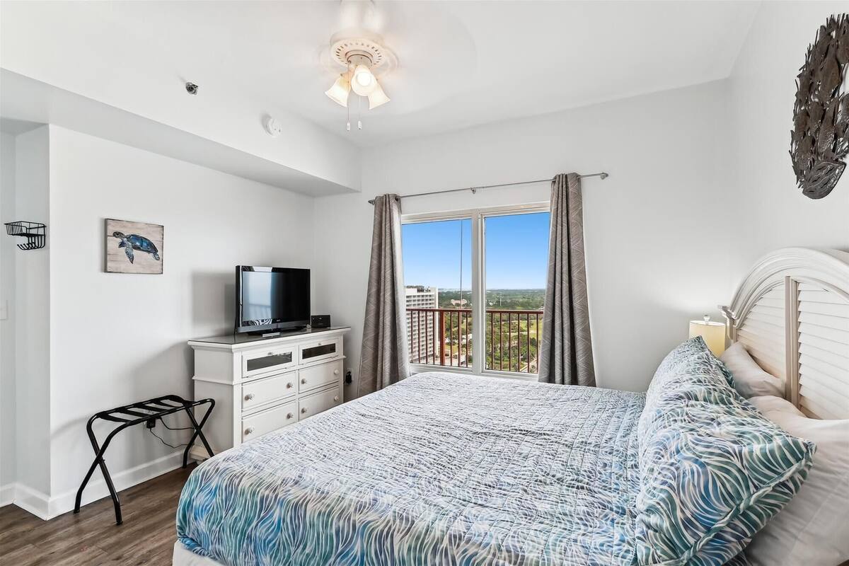 Ein Schlafzimmer mit Bett, Fernseher auf einer Kommode, einem Fenster mit Vorhängen und Balkonblick.