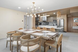 A modern kitchen with stainless steel appliances, a dining area with a large table and wicker chairs, and a hanging light fixture. - Designer Beachfront Condo with Endless Ocean Views (South Padre Island)