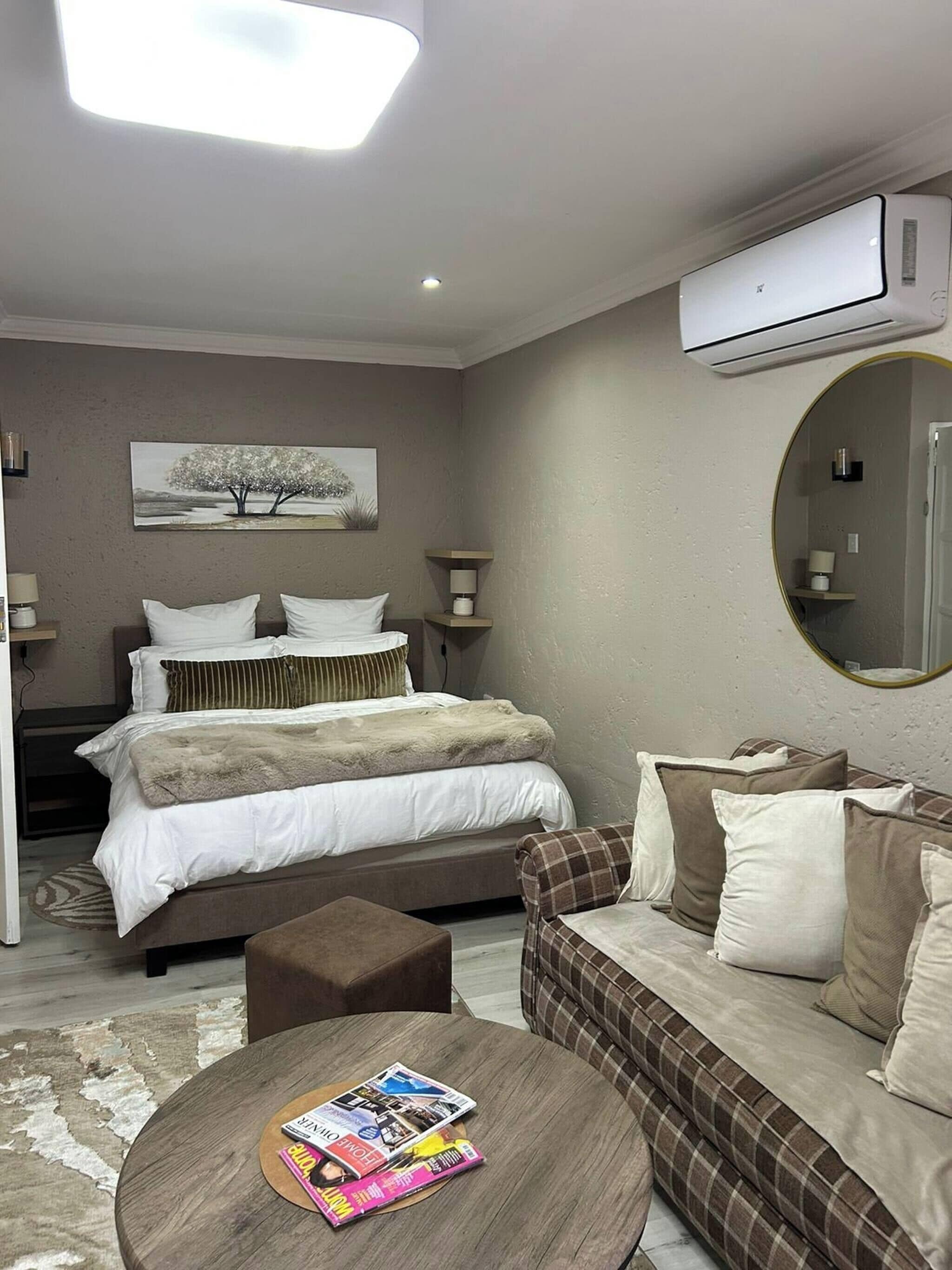A bedroom with a bed, a sofa, a coffee table with magazines, a painting on the wall, and an air conditioning unit.