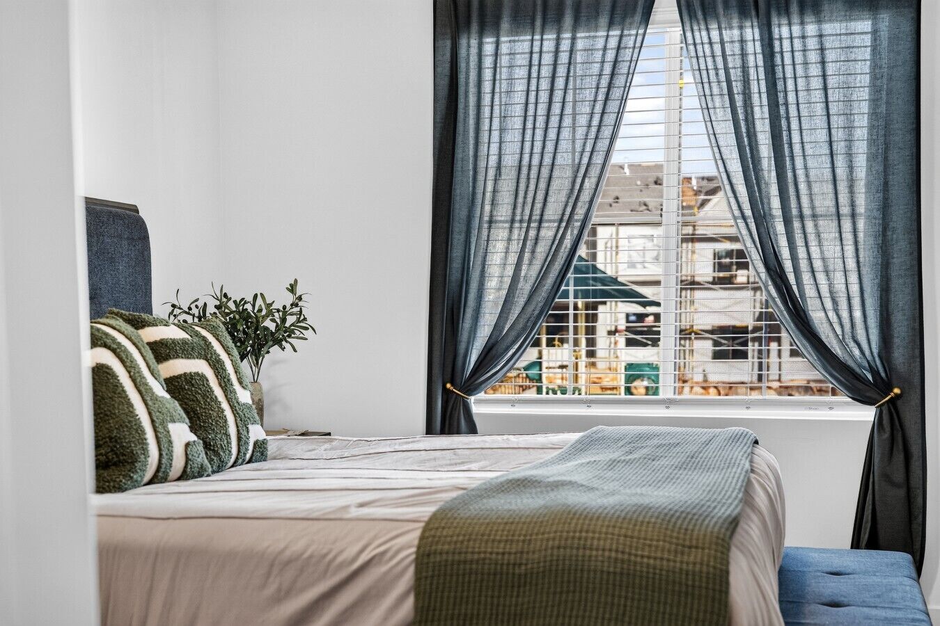 A bedroom with a bed, a window with curtains, a plant, and a view of buildings outside.