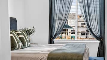 A bedroom with a bed, a window with curtains, a plant, and a view of buildings outside.