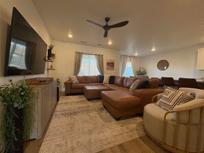 Ein Wohnzimmer mit Ecksofa, Couchtisch, Wand-Fernseher, Deckenventilator und Essbereich mit Stühlen.