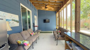 Un'area salotto esterna coperta con ventilatore a soffitto, televisore a parete e vista sul giardino.