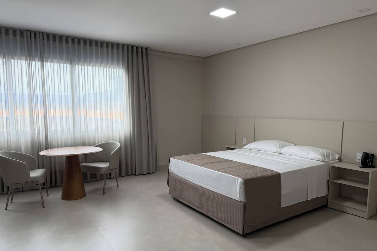 Quarto de hotel moderno com cama, mesa redonda, duas cadeiras, mesa de cabeceira e uma grande janela com cortinas.