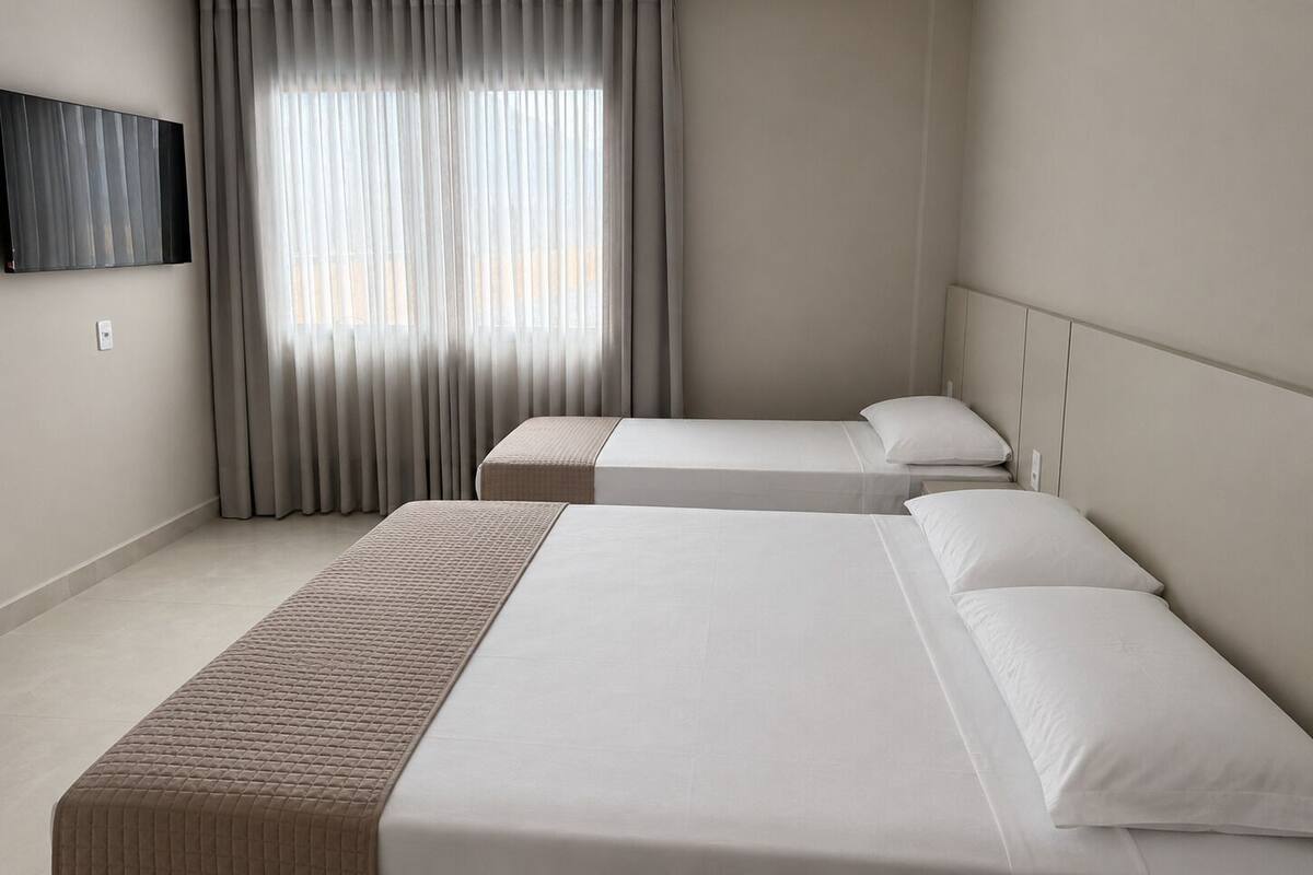 Quarto de hotel com duas camas, uma janela grande com cortinas, televisão de tela plana na parede e decoração em tons neutros.