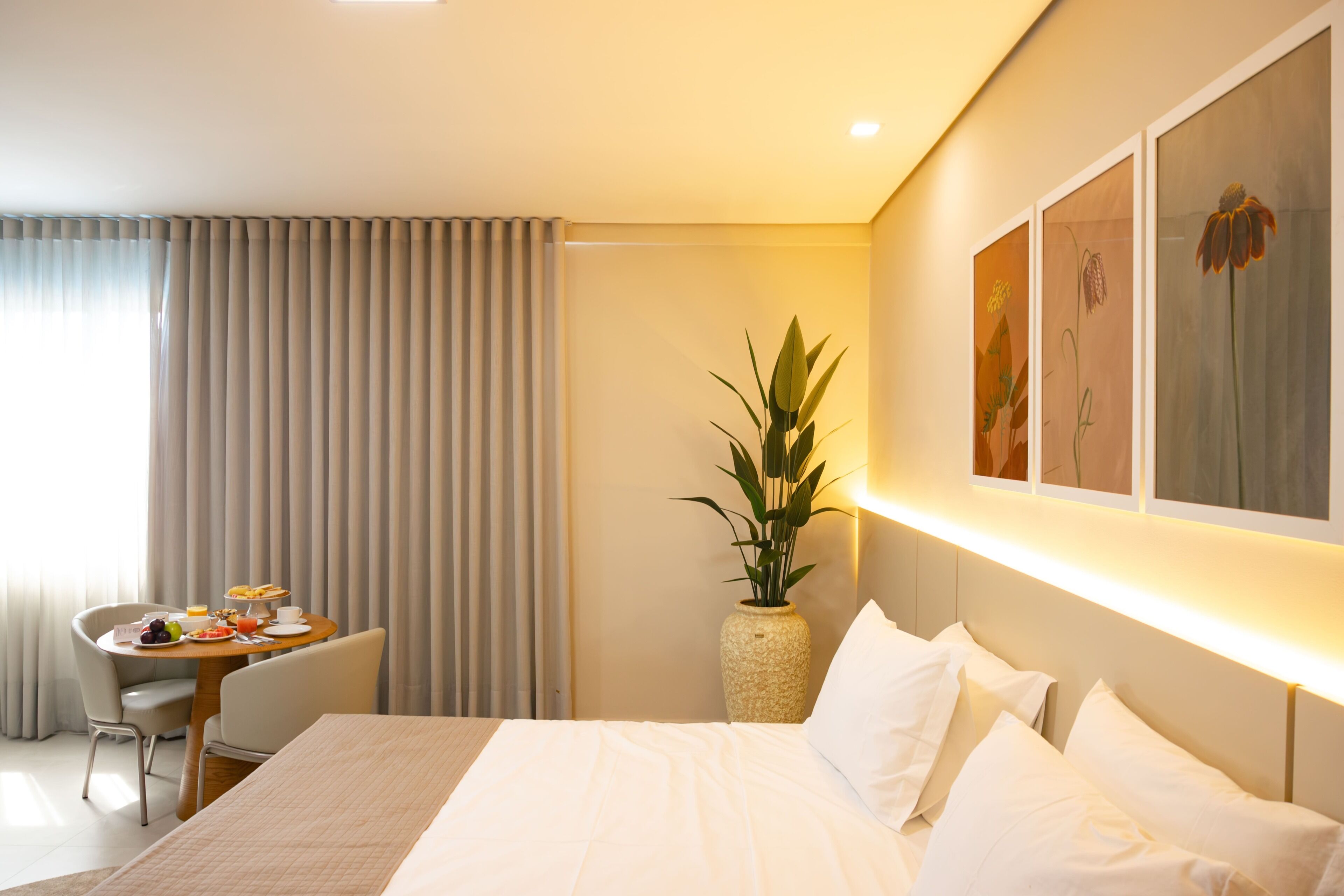 Ein Hotelzimmer mit einem Bett, einem Frühstückstisch, einer Pflanze und Kunstwerken an der Wand.