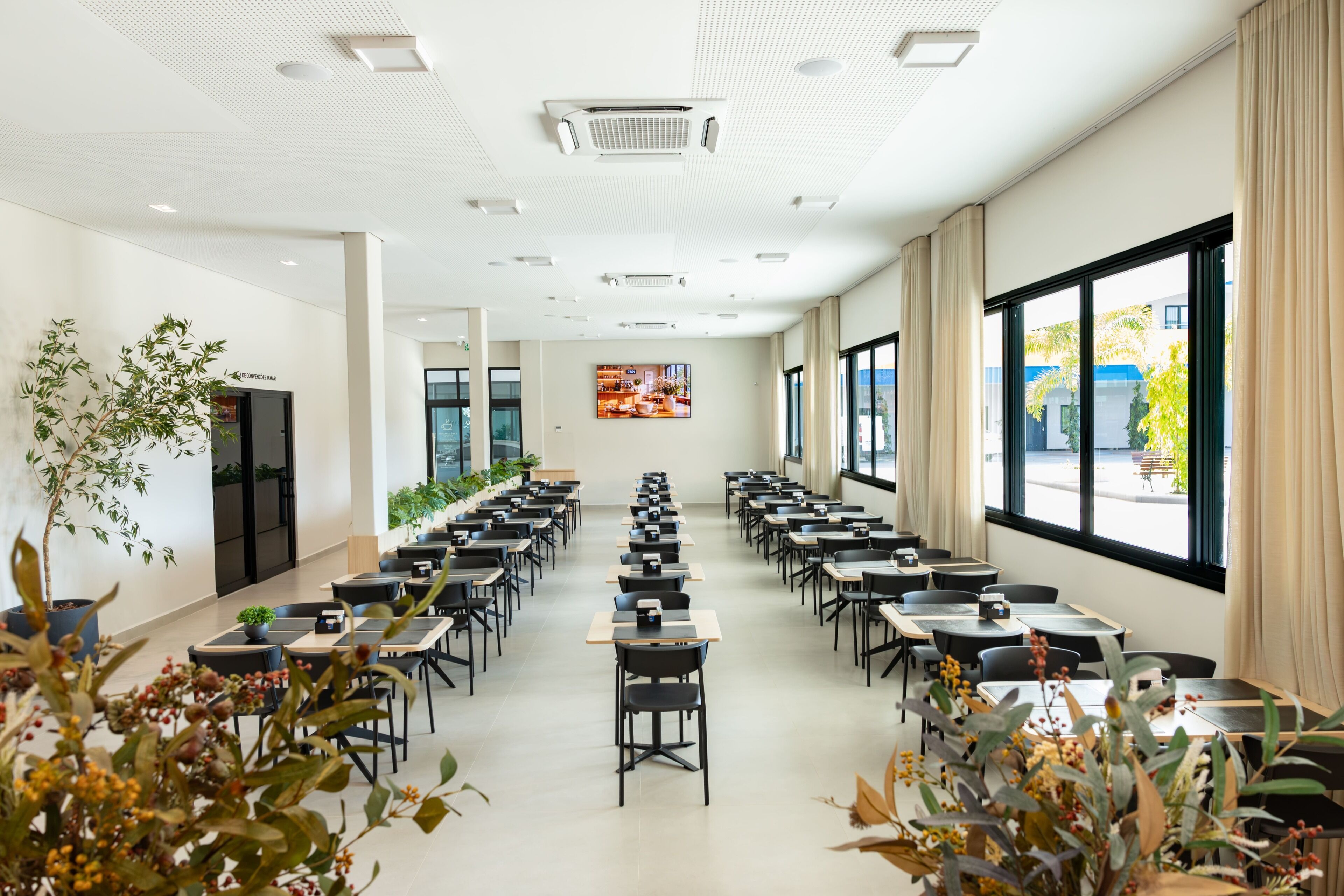 Un salón de conferencias con filas de mesas y sillas, ventanas amplias y luces en el techo.
