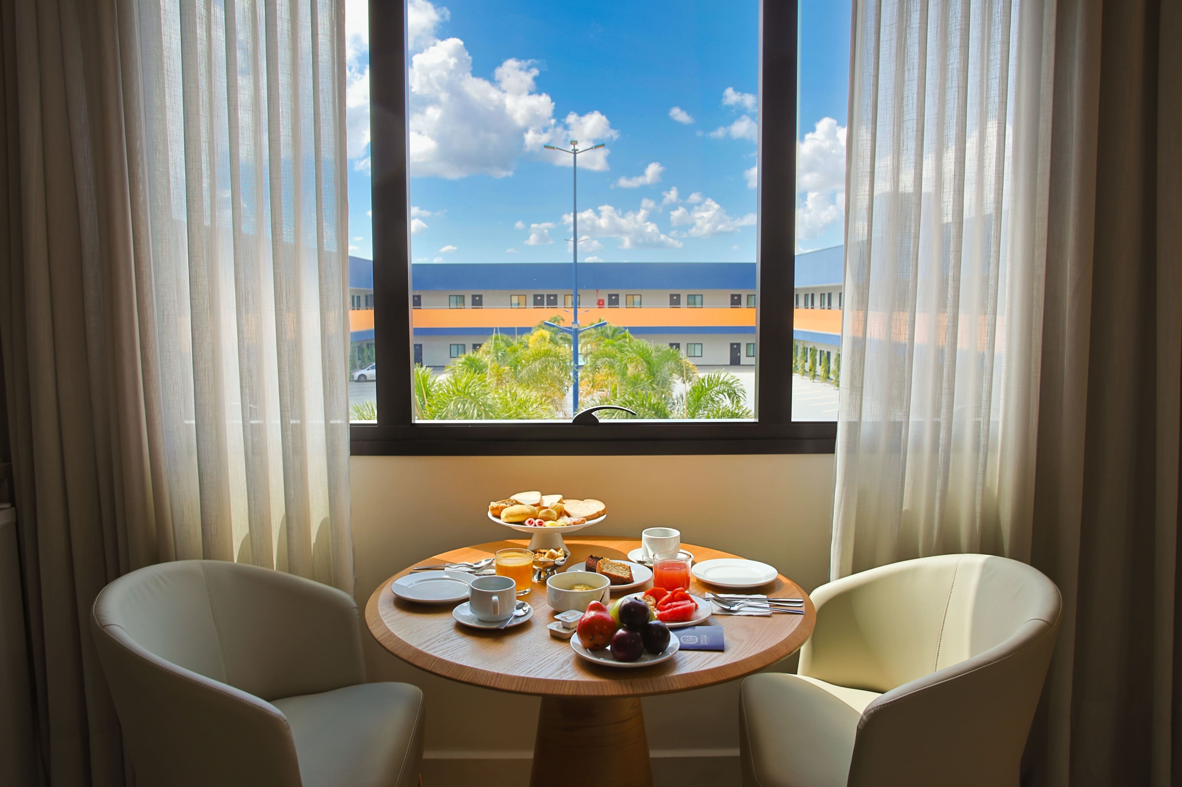 A dining area with a table set for breakfast, two chairs, and a view of a building through a window with curtains.
