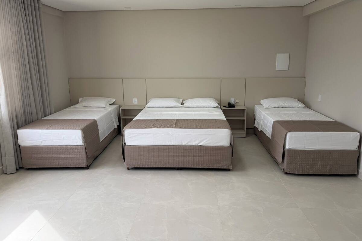 Quarto de hotel com três camas de solteiro, mesa de cabeceira com telefone e janela com cortinas.