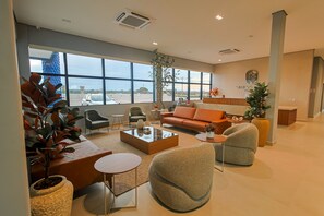 Un hall d’hôtel spacieux, doté d’une grande fenêtre offrant une vue sur l’extérieur. On y trouve plusieurs coins salon avec des canapés et des fauteuils, une table basse et des plantes en pot.