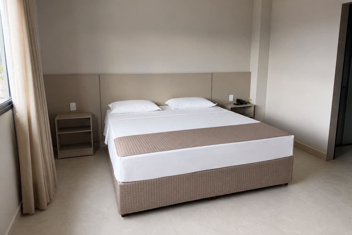 Quarto de hotel com uma cama grande, mesa de cabeceira e uma janela com cortinas.
