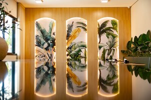 Un interior moderno con un mostrador de recepción de madera, un panel de vidrio con motivos de hojas tropicales y un reflejo en el suelo pulido.