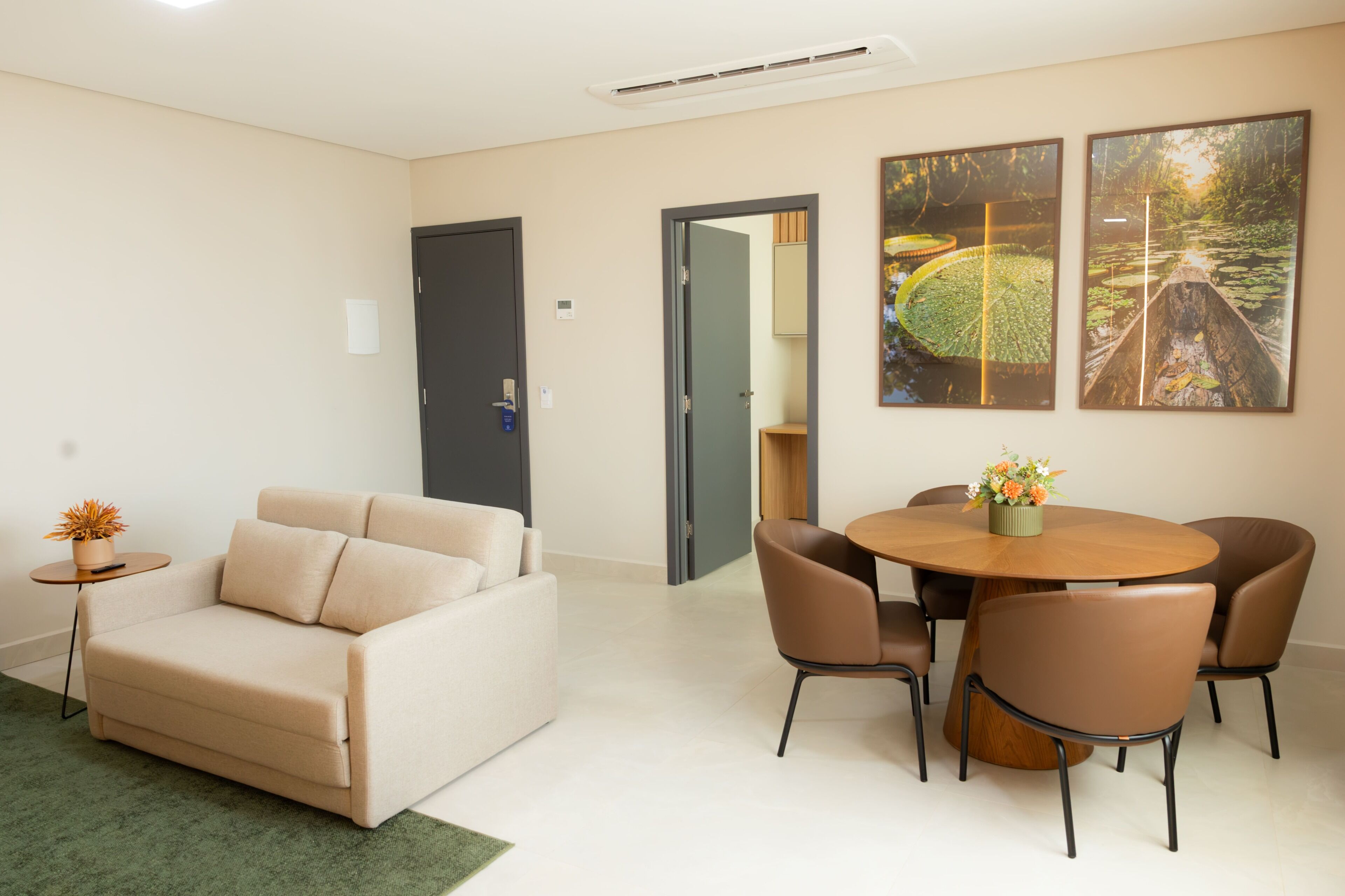 Uma sala de estar moderna com um sofá bege, uma mesa de jantar redonda de madeira com cadeiras e duas fotografias emolduradas da natureza na parede.