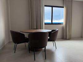 Uma área de jantar moderna com uma mesa redonda e quatro cadeiras, uma janela grande com vista para o céu e paredes em tons claros.