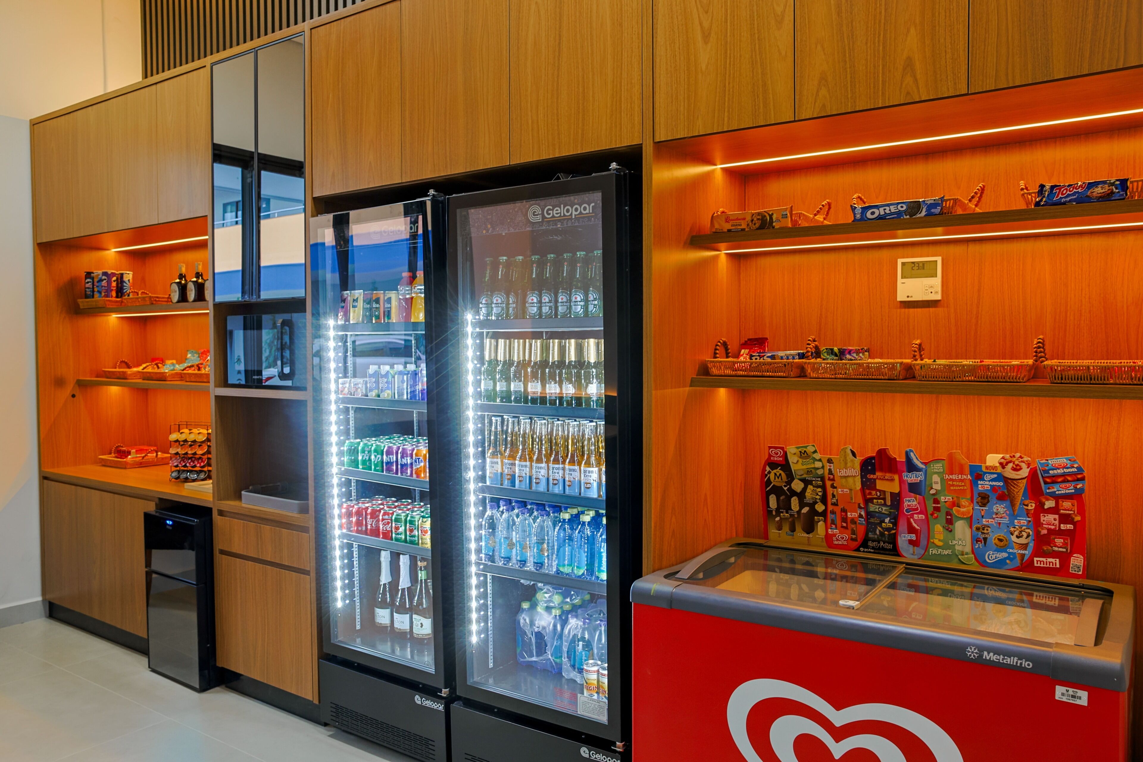 A hotel minibar with a variety of beverages and snacks, including a refrigerator stocked with bottles and cans, and shelves with packaged goods.