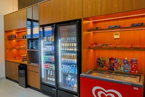 A hotel minibar with a variety of beverages and snacks, including a refrigerator stocked with bottles and cans, and shelves with packaged goods.