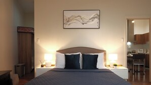 A neatly made bed with a dark blue quilt, two bedside lamps, a wall-mounted abstract art piece, and a visible kitchen area in the background. - New! 2BR w/ City Views in Makati & Fast Wi-Fi! (Makati)