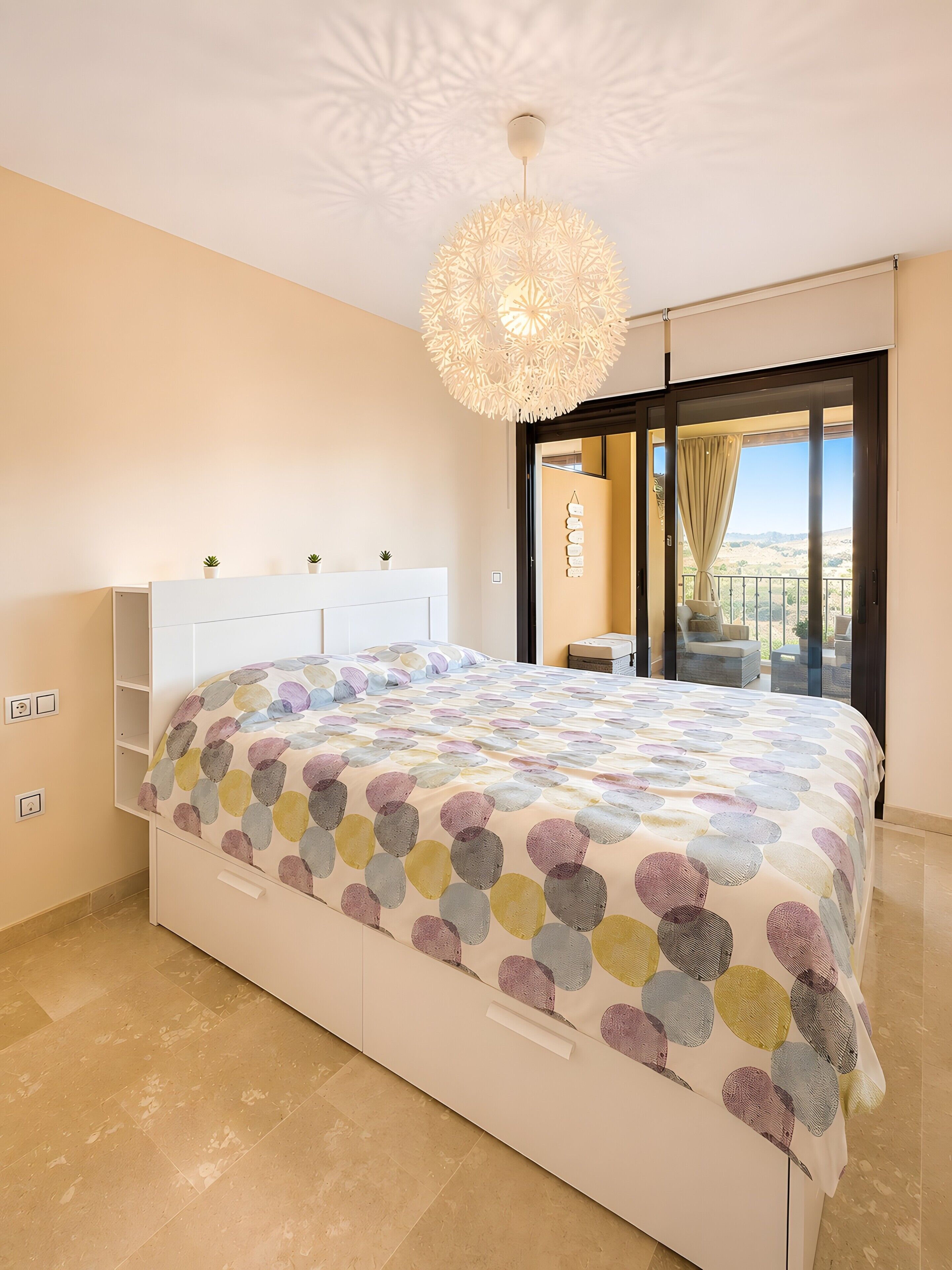 A bedroom with a bed, a ceiling light fixture, a sliding glass door to a balcony, and a wall-mounted power outlet.