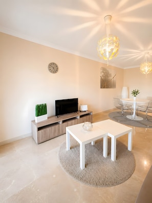 A modern living room with a flat-screen TV, a white coffee table, and a dining area with a round table and chairs.