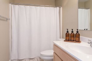 Ein Badezimmer mit weißer Duschvorhang, Toilette und Waschbecken. Auf dem Waschbecken stehen drei braune Flaschen.