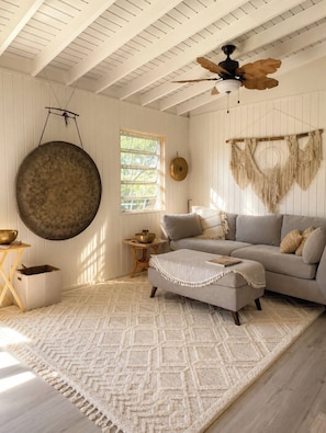 Um quarto com ventilador de teto, um painel redondo na parede, um painel de macramé, um sofá, um banquinho, uma mesinha com uma tigela e um tapete estampado.