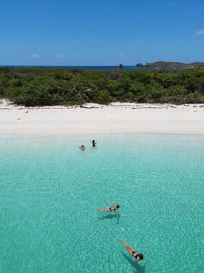 Een strand met helder turquoise water, een witte zandkust en weelderige groene begroeiing op de achtergrond.