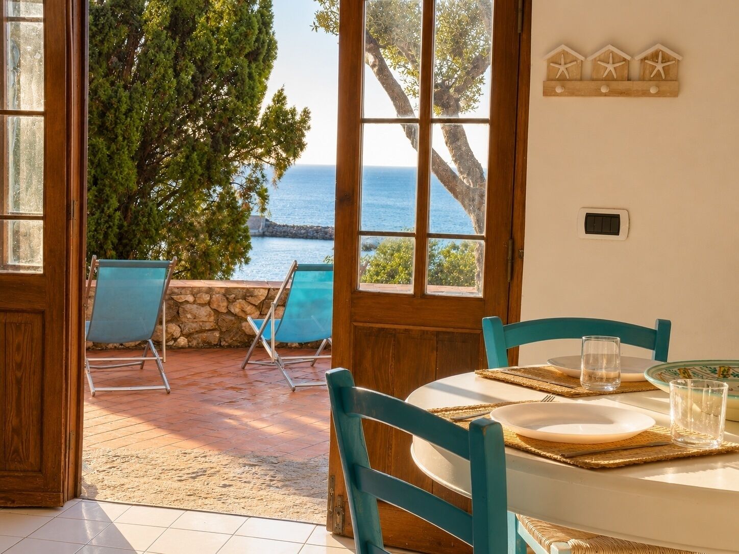 A room with a view of the sea, a dining table set with plates and glasses, and two blue chairs.