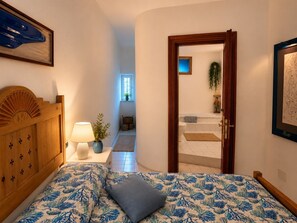 Ein Schlafzimmer mit einem Bett, einem Holzkopfende, einer Nachttischlampe, einer Topfpflanze und einem durchgegangene Tür erkennbaren Badezimmer.