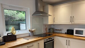 A modern kitchen with white cabinets, a built-in oven, a microwave, and a window with a view of greenery. - Cosy Cottage Retreat in Edinbane (Skeabost)