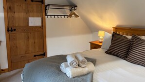 A bedroom with a wooden door, a bed with a grey blanket and rolled towels, a bedside table with a lamp, and a hanging light fixture. - Cosy Cottage Retreat in Edinbane (Skeabost)