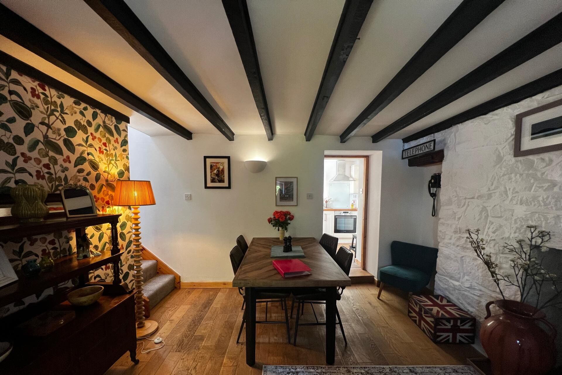 Un coin repas avec une table en bois, des chaises et un banc. Une lampe se trouve à gauche, et un vase avec des fleurs est posé sur la table. La pièce est dotée de poutres apparentes au plafond et d’un mur en pierre sur le côté gauche.