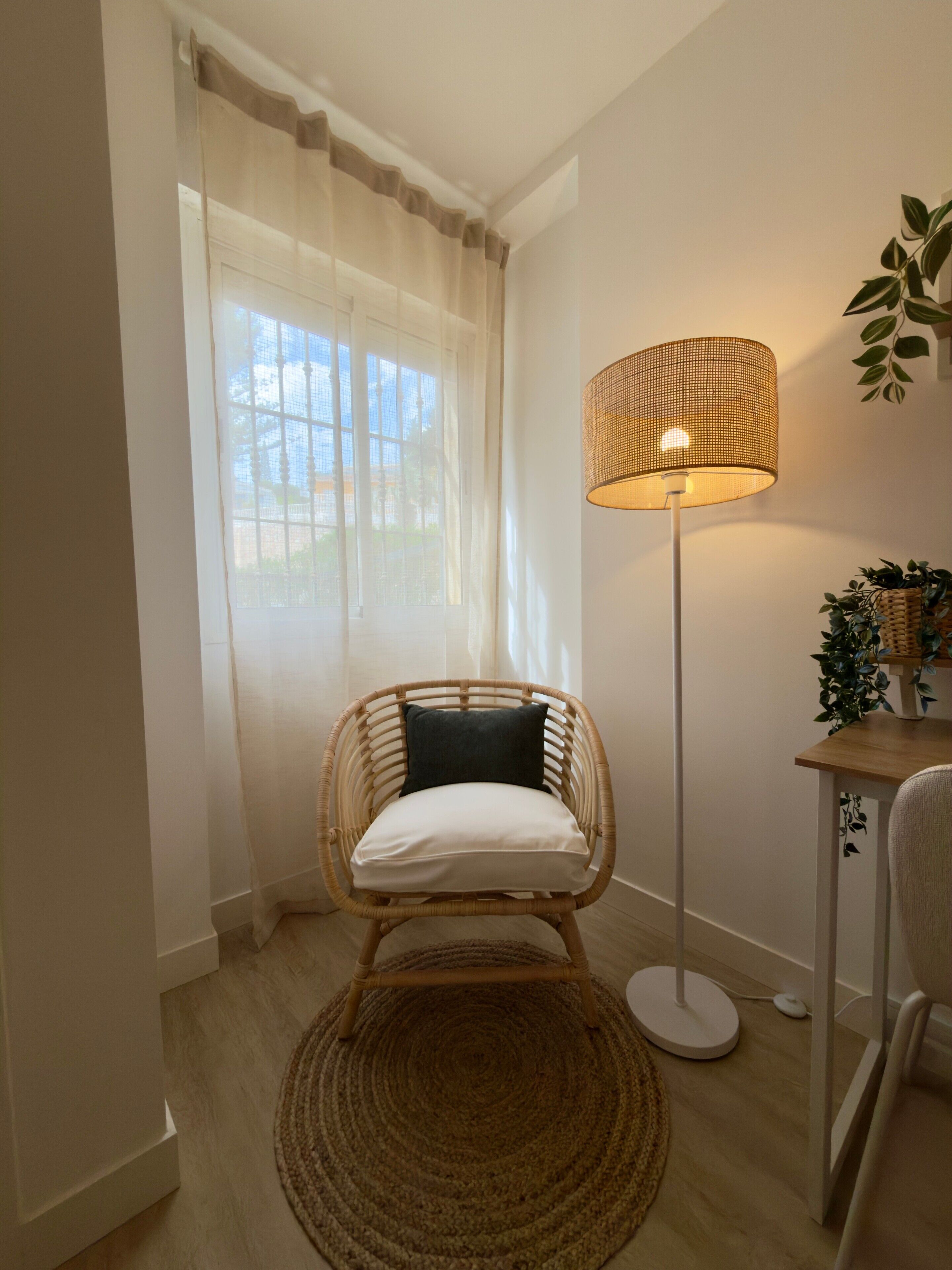 A room with a wicker chair, a floor lamp, a small table with a plant, and a window with sheer curtains.