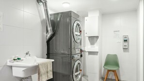 A compact bathroom with a grey wall-mounted washing machine, a white sink, a green chair, and a wall-mounted control panel.