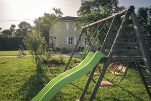 A playground with a slide and swings, a house in the background, and a person riding a bicycle. - Maison Bordeneuve: Sharing the Charm of the Gers (Préneron)