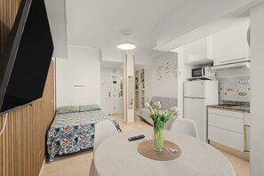 Un espace de vie compact comprenant un lit, une table à manger agrémentée d’un vase de fleurs, un coin cuisine avec un four à micro-ondes et un réfrigérateur, et un téléviseur fixé au mur.