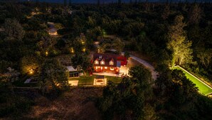 A large house surrounded by dense forest, with a well-lit driveway and a tennis court. - Luxury Retreat| Theater, Golf, Spa, Firepit & Views (Ahwahnee)