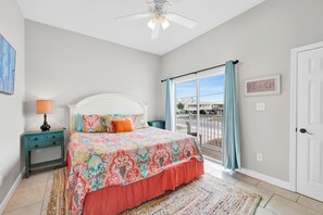 A bedroom with a bed, a nightstand, a lamp, a ceiling fan, a window with curtains, and a door. - Dolphin View Beachfront Duplex B | Pet Friendly (Gulf Shores)