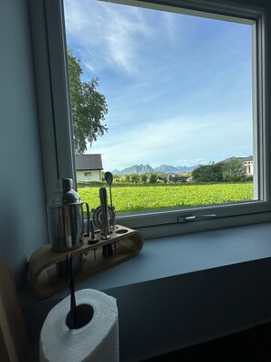 Une fenêtre donnant sur un champ verdoyant, un bâtiment et des montagnes au loin.