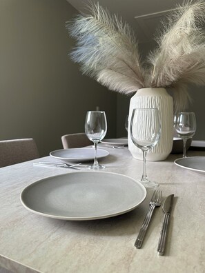 Une table à manger dressée avec des assiettes, des couverts et des verres à vin, agrémentée d’un vase contenant des plumes d’herbe des pampas.
