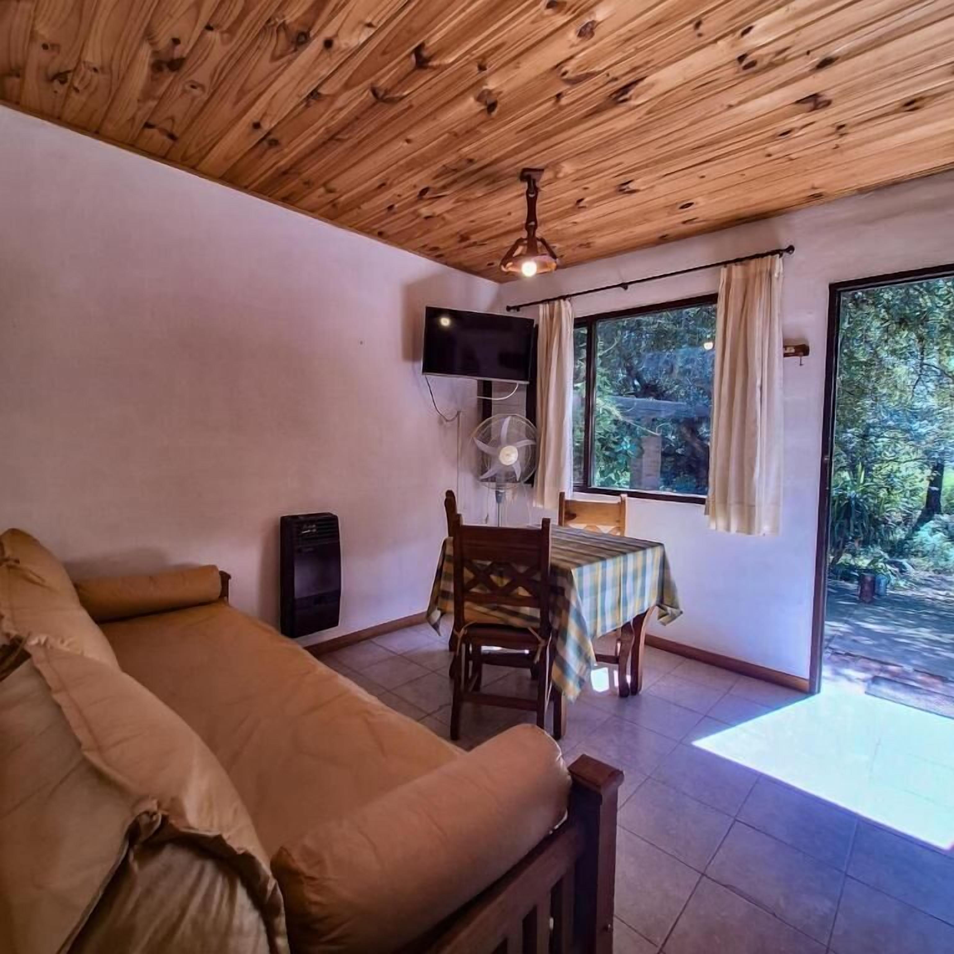 A room with a wooden ceiling, a bed, a TV, a table with a chair, and a window with curtains.