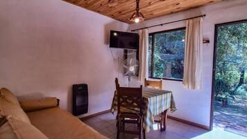 A room with a wooden ceiling, a bed, a TV, a table with a chair, and a window with curtains.