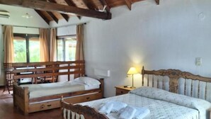 Una habitación con dos camas de madera, un banco de madera, una mesita de noche con lámpara y una ventana con cortinas.