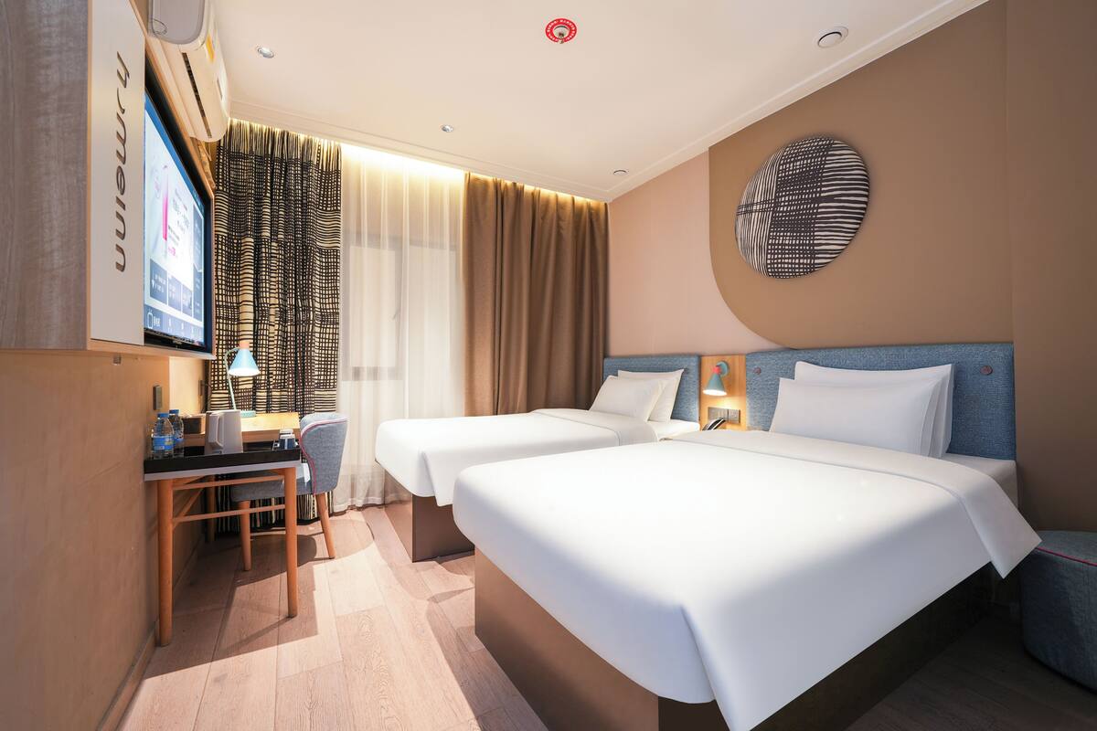 Una camera d'albergo con due letti, una scrivania, una sedia, una TV e un'opera d'arte appesa al muro.