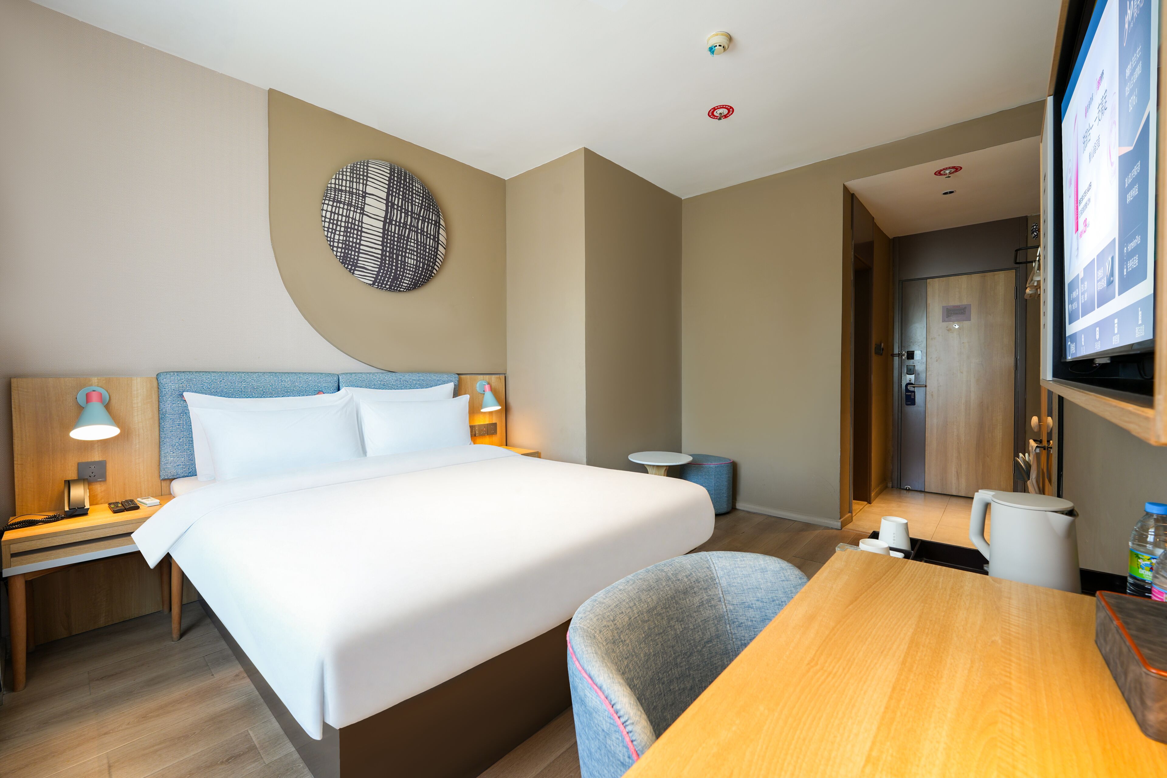 Une chambre d’hôtel comprenant un lit, des tables de chevet, un bureau, une chaise, un téléviseur et un petit élément de décoration circulaire fixé au mur.