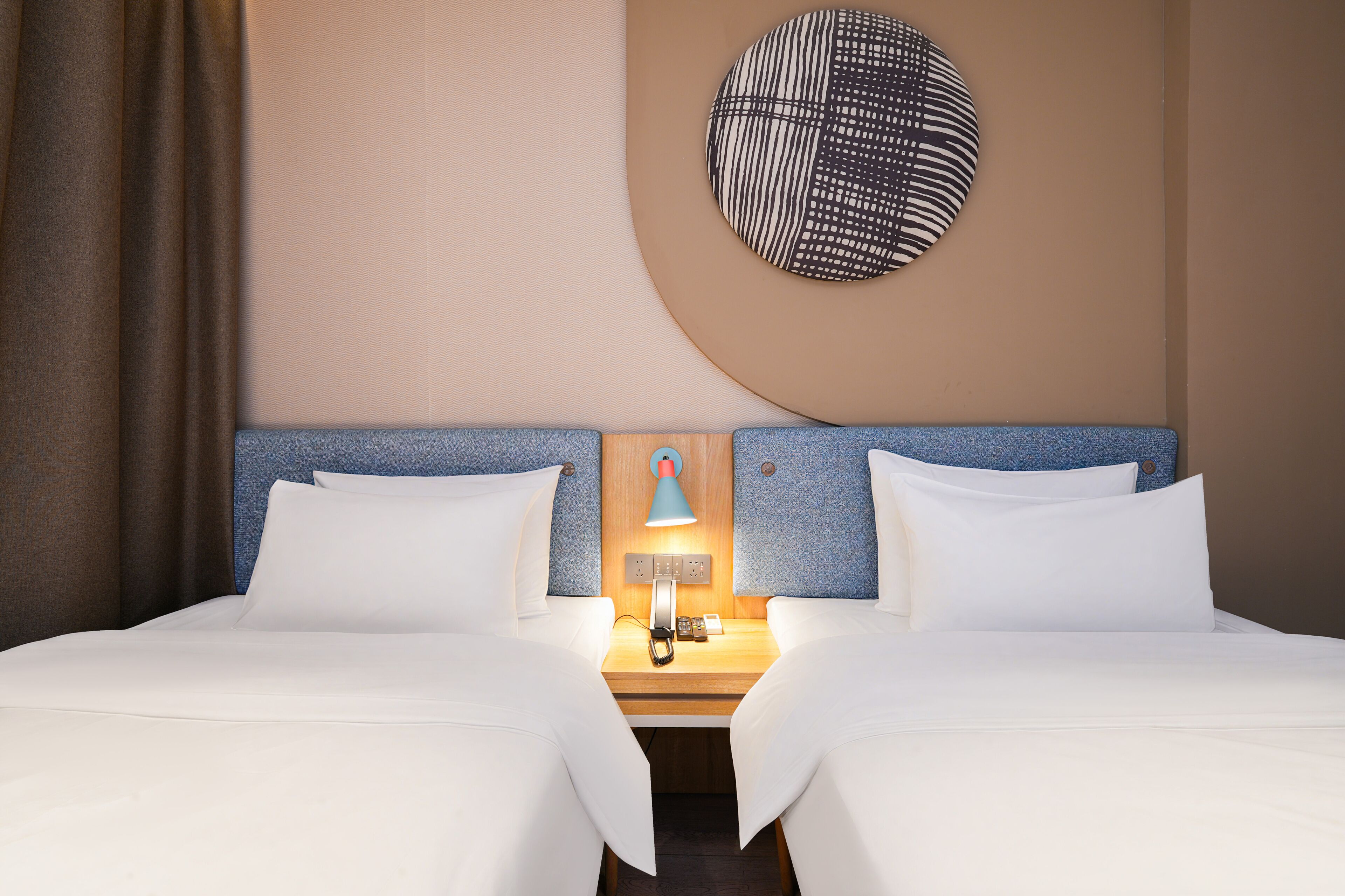 Quarto de hotel com duas camas, uma mesa de cabeceira com abajur e uma obra de arte circular pendurada na parede.