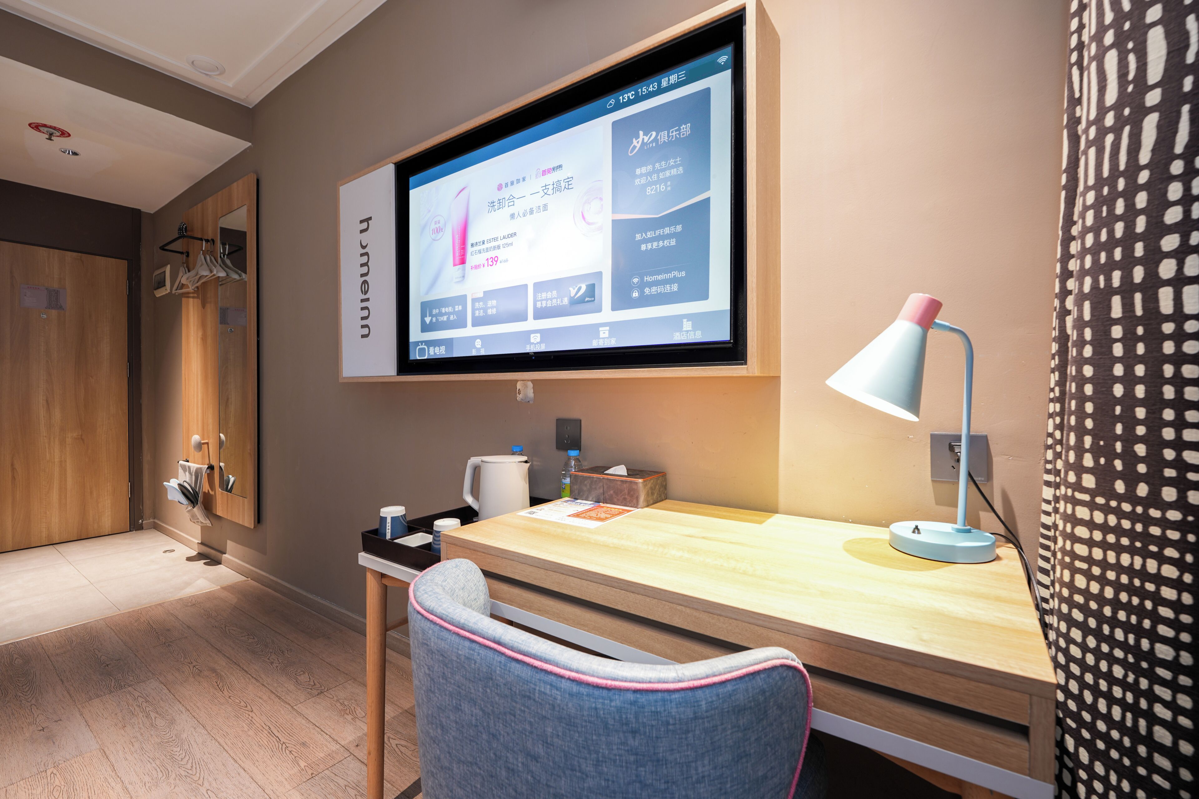 Een hotelkamer met een houten bureau, een stoel, een flatscreen televisie aan de muur, een lamp en een bed met een gordijn met patroon.