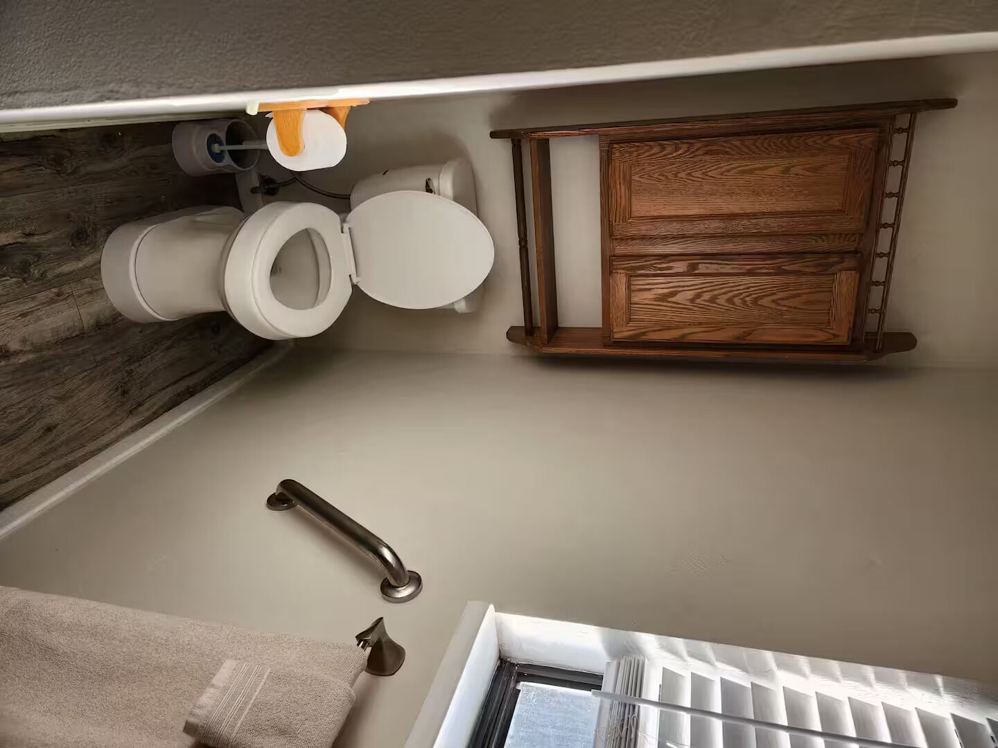 A bathroom with two toilets, a wooden cabinet, a handrail, and a window.