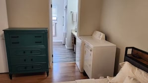 Een slaapkamer met een bed, een dressoir, een wasmachine en een gang naar een badkamer.