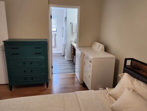 Ein Schlafzimmer mit Bett, Kommode, Waschmaschine und einem zur Toilette führenden Gang.